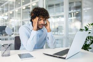 A young man experiences stress while working on his laptop at a modern, well-lit office. He is displaying signs of headache or concentration difficulty. photo