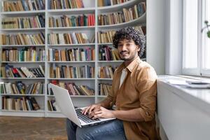 An engaged young adult sits comfortably in a well-lit space, surrounded by books, focused on work being done on a laptop. Setting suggests a modern library or a study room with a peaceful atmosphere. photo
