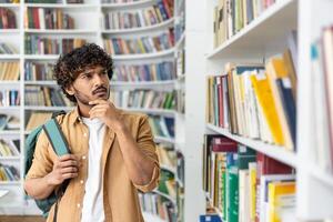 Thoughtful arab customer with backpack on one shoulder holding chin with hand while passing by bookcase. Worried man looking at rows of colourful encyclopaedias and looking for necessary dictionary. photo