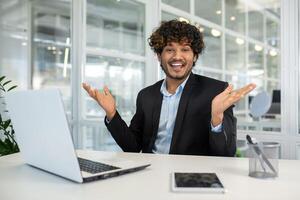 Cheerful young businessman with curly hair shrugs humorously during a presentation in a bright, modern office setting. Emphasizing a light-hearted approach to business communications. photo