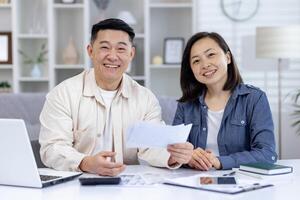 Happy asian couple sitting at table managing their finances using a laptop and documents, showing a positive family bond. photo