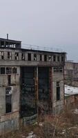 Old abandoned plant with broken windows ruined after the military actions. Demolished plant with pitted roof and some snow on the ground in winter. Aerial view Vertical video