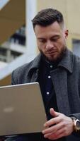 retrato do uma bonito homem com uma computador portátil. jovem barbudo homem de negocios dentro uma casaco trabalho em uma sem fio gadget em a urbano fundo ao ar livre. vertical video