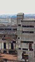 A huge state-owned factory with gray walls and broken glasses in the windows on the background of city buildings. Industrial zone. Aerial view. Vertical video