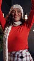 Beautiful girl with Christmas presents indoors. Portrait of a happy young woman wearing red sweater, knitted cap and scarf throws gift on Christmas background at home. Vertical video
