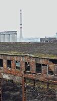 An old abandoned factory hangar with damaged walls on the background of other old structures in the same area. Aerial view. Vertical video