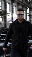 Strong man with a bat in abandoned place. Stylish man in glasses and black wearing moving forward on expensive car background inside the old empty plant. Vertical video