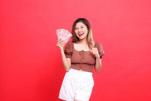 The expression of a cheerful Asian woman holding rupiah money while clenching her fists in front of her, wearing a brown blouse with a red background. for transaction, technology and advertising photo