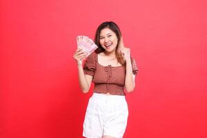 gesture of a cheerful indonesia woman clenching her fist successfully while holding rupiah money wearing a brown blouse top with a red background. for transaction, technology and advertising concepts photo