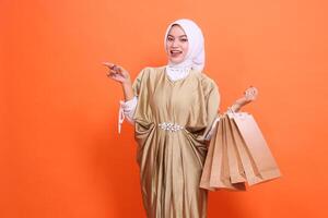 mature Asian woman is happy carrying a brown paper bag containing shopping items while pointing right wearing a kaftan Muslim dress with a hijab. Concept of fashion, transaction, promotion photo