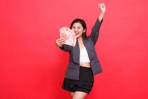 The expression of a cheerful Asian leader woman holds a credit card and money in front of her, clenches her fists and wears a jacket and skirt on a red background. for business and advertising photo