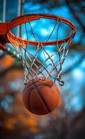 A basketball is suspended in the air above a basketball net. The ball is in the middle of the net and is surrounded by the net's strings photo