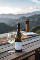 White wine in a bottle with a blank mock-up label and in a glass on a wooden outdoor table overlooking the mountains photo