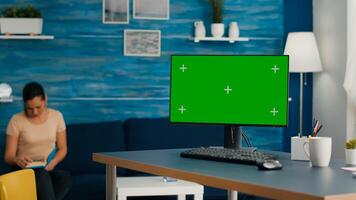Powerful computer with mock up green screen chroma key stands on office desk in living room. Woman reading book in background in bright room with Pc isolated display photo