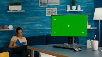 Woman lying on sofa reading a book in living room equipped with desktop computer with mock up green screen chroma key standing on table. Pc with isolated display is ready for online business photo
