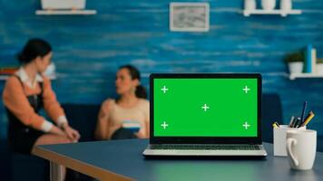 Two women sitting together on sofa in background talking about internet technology. In front on desk table standing isolated laptop computer with mock up green screen chroma key in office home photo