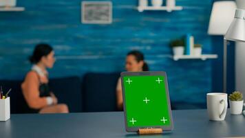 Isolated tablet computer with mock up green screen chroma key standing on desk table in living room. Two friends sitting on sofa in background talking about communication photo