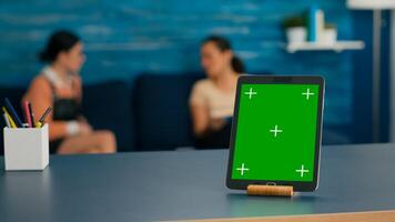 Two women sitting on sofa talking about digital technology. Office room is equipped with isolated tablet computer with mock up green screen chroma key display standing on table desk photo