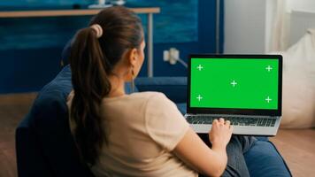 Freelancer woman sitting on sofa in home office typing on laptop computer with mock up green screen chroma key display. Caucasian woman working on financial graphs using isolated pc photo