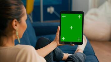 Caucasian female lying on sofa having online meeting on tablet computer with mock up green screen chroma key display. Woman using isolated touchscreen device for social networks browsing photo