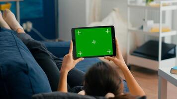 Woman sitting on sofa in living room watching movies on tablet computer with mock up green screen chroma key display. Freelancer using isolated touchscreen device for social networks browsing photo