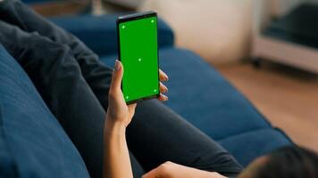 Freelancer holding in vertical mode smartphone with mock up green screen chroma key display while lying on sofa in living room. Woman using isolated touchscreen device for social networks browsing photo