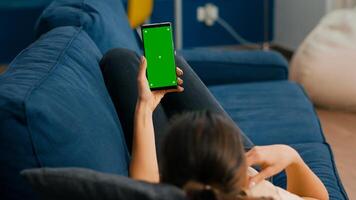 Freelancer lying on sofa while having online videocall on smartphone with mock up green screen chroma key display in vertical mode. Woman using isolated touchscreen device for social networks browsing photo