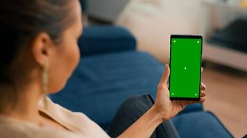 Caucasian female sitting on sofa in living room while socializes on internet app using phone with mock up green screen chroma key display.Woman browsing on network with isolated gadget photo