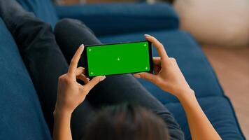 Freelancer sitting on sofa while looking at movies using phone in horizontal mode with mock up green screen chroma key display. Woman using isolated touchscreen device for social networks browsing photo