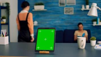 Home studio equipped with tablet computer with mock up green screen chroma key display standing in vertical mode on office desk. In background two collegues talking about internet advertising photo