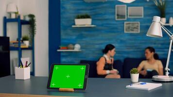 Tablet computer with mock up green screen chroma key display standing in front on office desk. Two collegues talking in background, sitting on sofa about internet connection photo