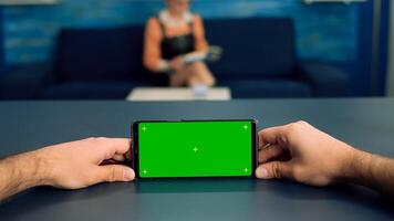 Pov of man hands holding in horizontal mode smartphone with mock up green screen chroma key display standing on desk in office studio. Freelancer using isolated phone for browsing social networks photo