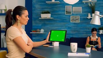 Woman watching online videos on tablet computer with mock up green screen display while sitting at office desk. Freelancer looking at template chroma key isolated device using technology internet photo