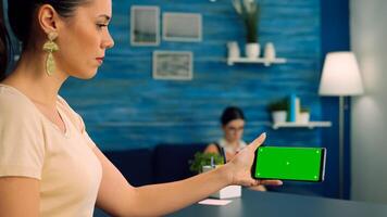 Caucasian female holding isolated smartphone with mock up green screen chroma key display, browsing online information on internet while her collegue reading a book in background photo