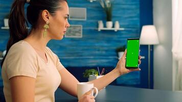Casual woman having videocall with her collegue while drinking cup of coffee using mock up green screen chroma key phone. Caucasian female working on social media project sitting on office desk photo