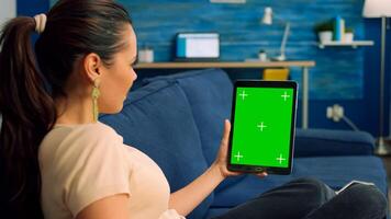 Freelancer woman looking at green mock up screen tablet computer in vertical portrait mode while standing on couch. Caucasian female watching online videos using isolated device photo