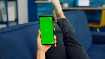 Freelancer woman lying on sofa looking at mock up green screen chroma key smartphone in vertical mode. Caucasian female browsing on internet using isolated phone for social media network photo