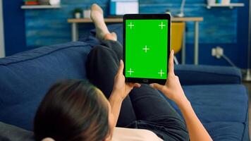Casual woman lying on sofa using tablet computer mock up green screen chroma key in vertical portrait mode. Freelancer browsing on internet with isolated device for online project photo