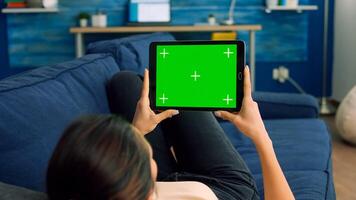 Over the shoulder view of casual woman sitting on sofa using tablet computer with mock up green screen chroma key. Caucasian female browsing on internet use touchscreen device photo