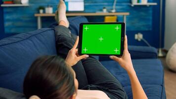 View of shoulder of woman uses isolated tablet computer with mock up green screen chroma key sitting on sofa in living room. Freelancer using technology communication concept for online template photo