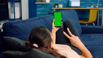 Casual woman holding smartphone with mock up green screen chroma key during online videocall while lying on sofa. Freelancer using technology communication concept for online template photo
