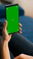 Freelancer holding in vertical mode smartphone with mock up green screen chroma key display while lying on sofa in living room. Woman using isolated touchscreen device for social networks browsing photo