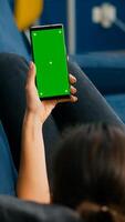 Freelancer lying on sofa while having online videocall on smartphone with mock up green screen chroma key display in vertical mode. Woman using isolated touchscreen device for social networks browsing photo