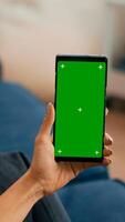 Caucasian female sitting on sofa in living room while socializes on internet app using phone with mock up green screen chroma key display.Woman browsing on network with isolated gadget photo