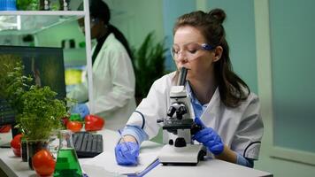 Biologist scientist looking at leaf sample using medical microscope while writing gmo expertise on notepad. Chemist examining biological discovery on plant working in pharmaceutical laboratory photo