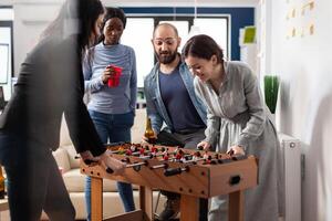 Multi ethnic group of people playing at foosball table after work partying and enjoying alcohol cups bottles of beer. Diverse team colleagues celebrating break from business activities photo