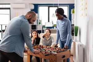 Multi ethnic friends enjoy after work foosball game at table soccer football while holding cups bottles of beer alcohol. Diverse group of colleagues having fun entertainment enjoyment photo