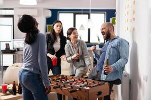 Multi ethnic friends enjoy after work foosball game at table soccer football while holding cups bottles of beer alcohol. Diverse group of colleagues having fun entertainment enjoyment photo