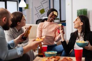 African american woman playing guessing game with friends after work at office. Multi ethnic group of workers guess imitation for fun cheerful activity while eating and drinking beer photo