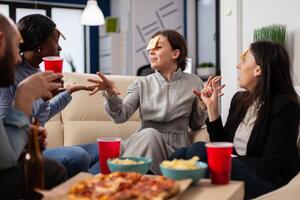 Diverse group of friends play guess who game after work while eating pizza chips and drinking beer. Multi ethnic colleagues enjoy fun cheerful activity at office with food on table photo